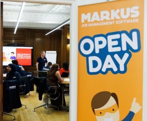 A photo looking through a door to a room full of people, poster to the right saying MARKUS OPEN DAY